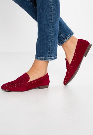 Kvinde iført blå jeans og røde ruskindsløfler med metalliske nitter, stående med den ene fod let hævet.