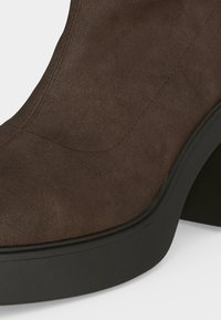 Gros plan d'une bottine en daim marron avec talon et semelle noirs épais, montrant les coutures détaillées sur la partie supérieure.