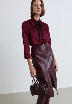 Donna che indossa una camicetta bordeaux con fiocco nero, gonna in pelle abbinata con spacco frontale, che tiene una borsa marrone scuro su sfondo uniforme.