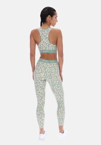 Femme aux cheveux tressés portant un soutien-gorge de sport et un legging à imprimé léopard vert et beige avec une ceinture "TED BAKER SPORT", debout de dos.