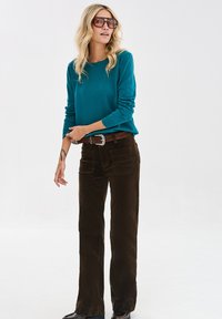 Kobieta z blond włosami, nosząca duże okulary przeciwsłoneczne, turkusowy sweter, brązowy pasek, ciemnobrązowe spodnie i czarne buty, stojąca na tle jednolitym.