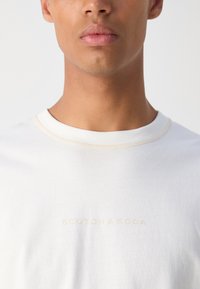 Hvid bomulds t-shirt med rund halsudskæring, prydet med diskret beige syning og et "Scotch & Soda" logo i lys beige på forsiden.