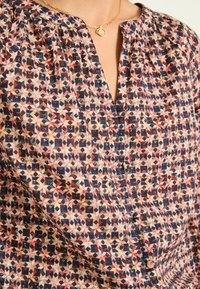 Multicolored blouse made of lightweight fabric, featuring a geometric pattern in pink, navy, and cream with a V-neckline and subtle pleats.