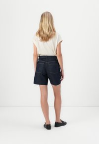 Mørkeblå denimshorts med rå kant, to baklommer, kombinert med en lys kremfarget kortermet topp og svarte flate sko.