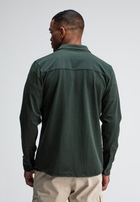 Mann, der von hinten schaut, trägt ein dunkelgrünes langärmliges Hemd und beige Hosen vor einem schlichten weißen Hintergrund.