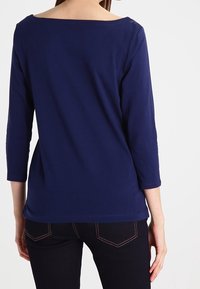 Kvinna i en marinblå långärmad skjorta och mörka jeans, sedd bakifrån mot en enkel vit bakgrund.