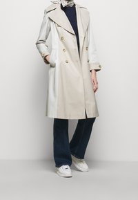 Persona que lleva un abrigo trinchera de color beige y blanco, jeans de color azul oscuro, zapatillas blancas y una bufanda blanca con patrón alrededor del cuello.