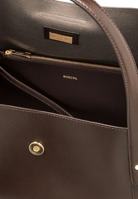 Brown leather handbag interior with a gold zip pocket and a gold brand plate. Features a circular magnetic snap and detailed stitching.