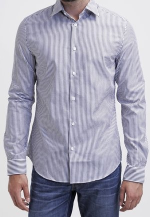 Mann trägt ein weißes Hemd mit langen Ärmeln und dünnen vertikalen blauen Streifen sowie eine blaue Jeans, steht mit entspannten Armen an den Seiten.