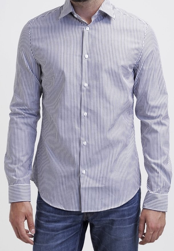 Homme portant une chemise blanche à manches longues avec de fines rayures verticales bleues et un jean bleu, debout les bras détendus le long du corps.