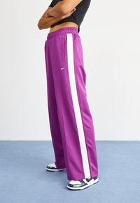 Purple athletic pants with white side stripes, elastic waistband, and pockets. Worn with black and white sneakers featuring reflective accents.