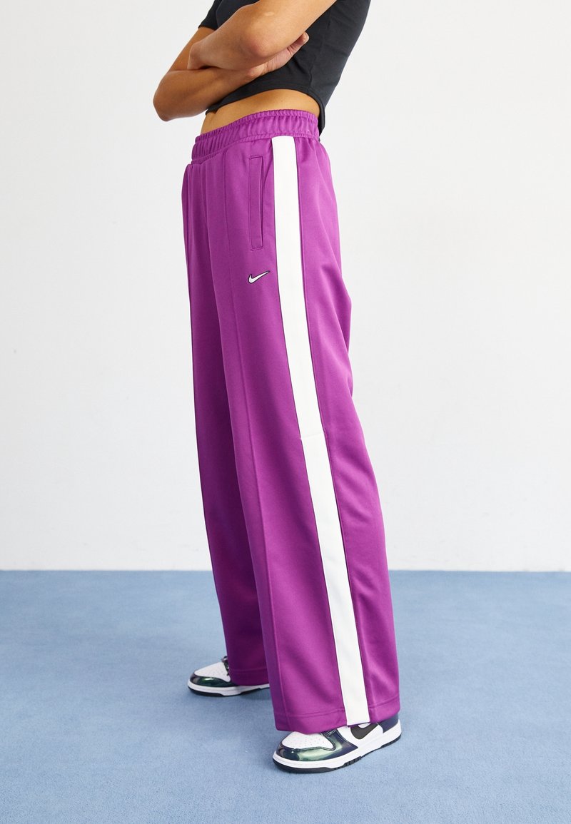 Purple athletic pants with white side stripes, elastic waistband, and pockets. Worn with black and white sneakers featuring reflective accents.