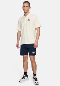 Crèmekleurig poloshirt met een klein rood en blauw logo, gecombineerd met marineblauwe shorts met witte tekst. Afgemaakt met zwart-witte sportieve sneakers.