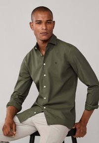 Olivengroen shirt met lange mouwen, een klassieke kraag, een knoopsluiting aan de voorkant en opgerolde mouwen. Voorzien van een klein logo op de borst.