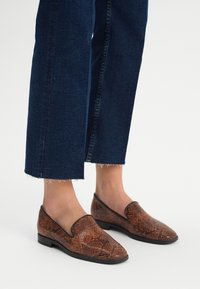 Bruine loafers met slangenprint, met een gladde textuur en een puntige neus, gecombineerd met donkerblauwe cropped jeans met rafelige zomen.