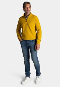 Geel, trui met ritssluiting in de hals, gecombineerd met blauwe jeans en witte sneakers. De trui heeft een zachte textuur; de jeans zijn aansluitend en licht beschadigd.