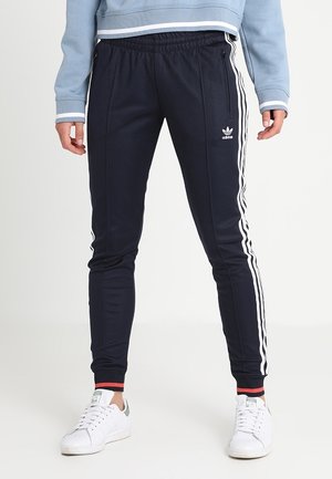Osoba nosząca czarne dresowe spodnie Adidas z białymi paskami i logo, w połączeniu z białymi sneakersami i niebieską bluzką z długim rękawem.