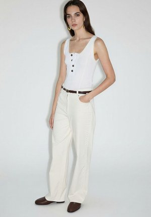 Woman wearing white sleeveless top with buttons, high-waist wide-leg cream pants, brown belt, brown shoes, and gold earrings, standing against white wall.