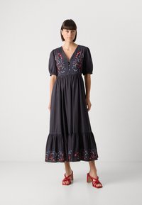 Dunkelblaues Baumwollkleid mit Puffärmeln und V-Ausschnitt. Verziert mit buntem Blumenstickerei und einem gestuften Saum. Getragen mit roten Absatzsandalen.