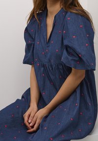 Denimimekko, jossa on lyhyet puff-hihat, v-pääkolo ja rypytetty vyötärö. Koko mekossa on pieniä punaisia sydänkuvioita. Pehmeä materiaali ja rento muotoilu.