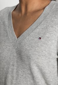 Grauer V-Ausschnitt-Pullover aus weichem Material, mit geripptem Kragen und einem kleinen, mehrfarbigen Logo auf der linken Brustseite.