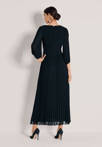 Zwarte geplooide maxi-jurk met een nauwsluitend lijfje, lange mouwen en een lage rug. Draagt hoge hakken en toont een vloeiende silhouet.