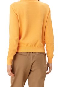 Orange strikpullover med rund halsudskæring og lange ærmer, der har ribbede manchetter og kant. Bæres med beige bukser.