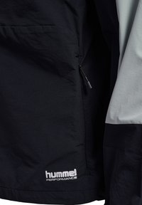 Mörkblå jacka med ljusgrå detaljer, utrustad med en dragkedjeficka och mjuk textur. "hummel PERFORMANCE"-logotyp i vitt på den nedre kanten.