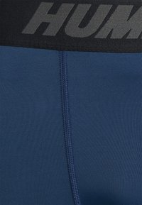 Marinblå idrottsshorts med en slät yta, med svart elastiskt midjeband med ordet "HUM" i fet grå typografi.