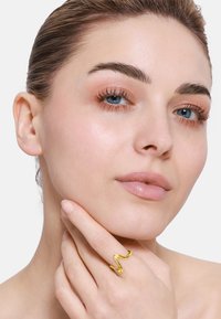 Anello zigzag in oro indossato su un dito, con una finitura lucida. La persona si tocca leggermente il viso, evidenziato da una pelle liscia.