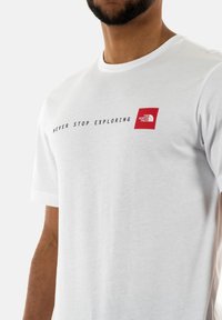 Camiseta de manga corta blanca con el texto "NUNCA DEJES DE EXPLORAR" en negro y un logotipo cuadrado rojo de The North Face en el lado izquierdo del pecho.
