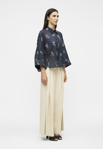 Femme debout portant une chemise ample à motif sombre avec de larges manches et un pantalon beige plissé à jambes larges avec des talons ouverts noirs.