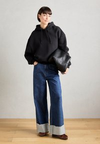 Black oversized hoodie, blue wide-leg jeans with frayed light denim cuffs, and brown patterned shoes, held with a black clutch bag.