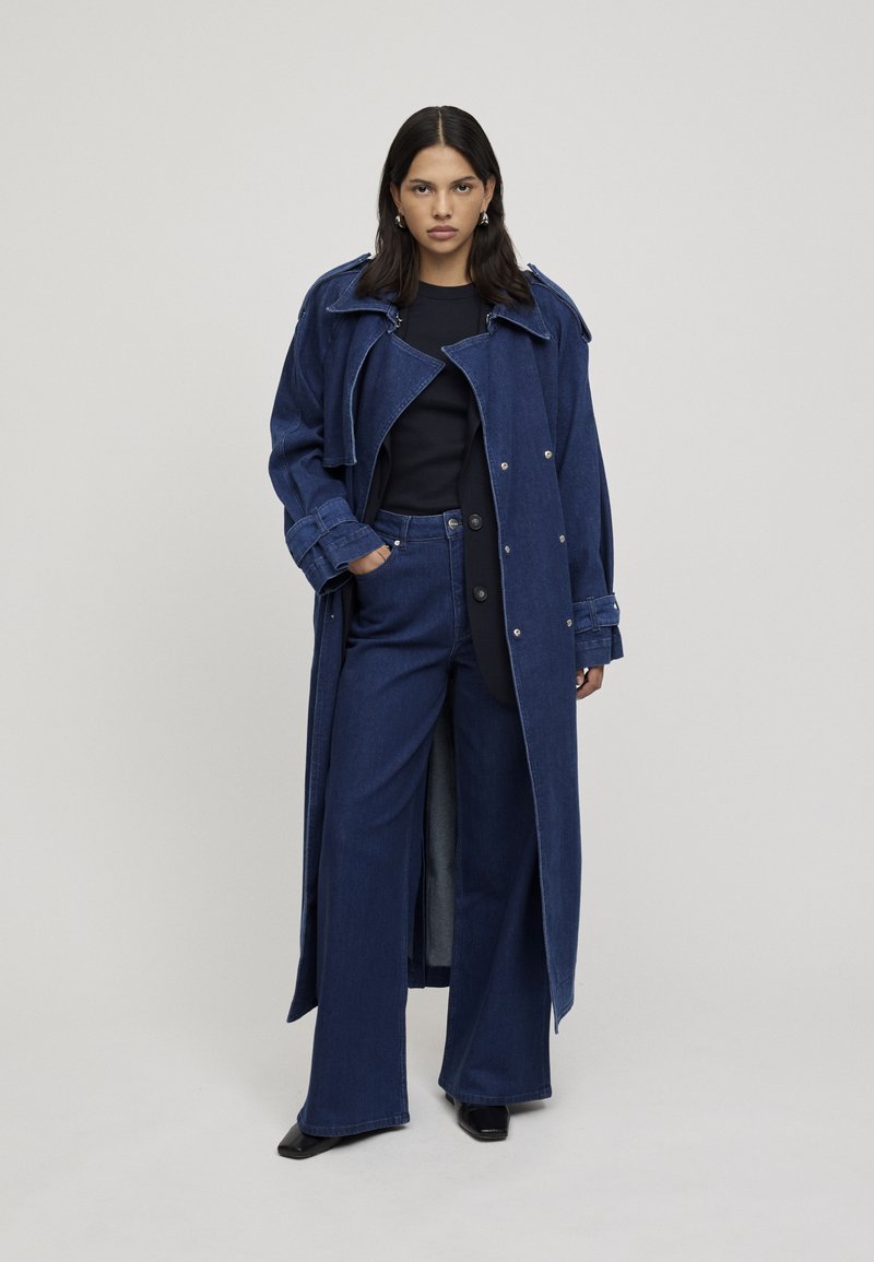 Lungo trench coat in denim blu con ampio colletto, bottoni grandi e spacchi laterali, abbinato a pantaloni in denim a gamba larga e un top nero.
