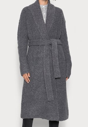 Kobieta ubrana w długi, teksturowany szary płaszcz z kołnierzem szalowym i paskiem do wiązania, na białej bluzce w wzory oraz czarnych spodniach.