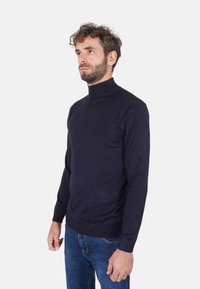 Maglione a collo alto blu navy realizzato in tessuto morbido e liscio. Design aderente con maniche lunghe e orlo a coste. Abbinato a jeans di denim.