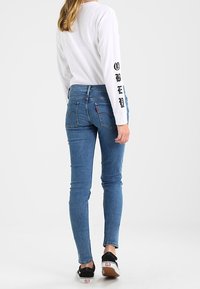 Jeans ajustados azules con cintura alta, bolsillos traseros detallados y una camiseta blanca de mangas largas con letras negras en la manga.