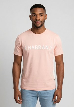 Joven con pelo corto y barba, vestido con una camiseta rosa claro con el texto "CHABRAND" y jeans azul claro, posando frente a un fondo liso.