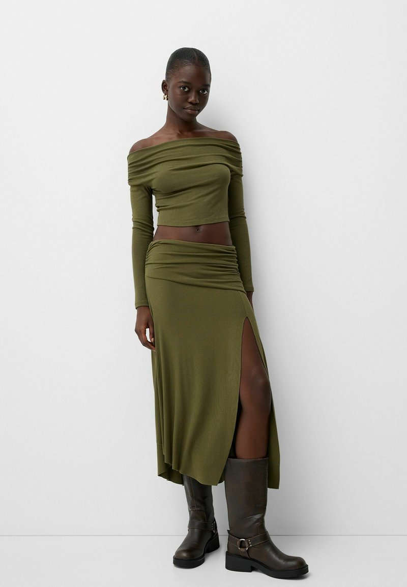Tenue deux pièces comprenant un haut olive dénudé sur les épaules et une jupe assortie avec une fente haute, complétée par des bottes marron montant jusqu'aux genoux.