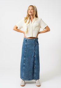 Chemisier court à manches courtes texturé blanc, jupe maxi en denim bleu taille haute avec détails de boutons, associé à des baskets légères et une écharpe à motifs.