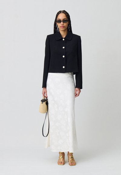 Chaqueta negra corta con grandes botones blancos, combinada con una falda maxi de patrón floral blanco y sandalias doradas de tiras. Pequeño bolso de mimbre.