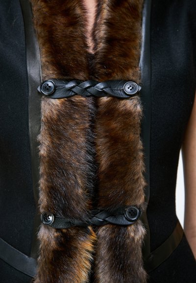 Gros plan d'une bordure en fourrure marron sur un vêtement noir maintenu par des lanières en cuir noir tressé et quatre boutons noirs.