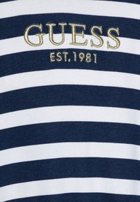 Majica s prugama u mornarsko plavoj i bijeloj boji s vezenim logom "GUESS" i "EST. 1981" u zlatnoj niti. Pamuk, kratki rukavi, okrugli izrez.
