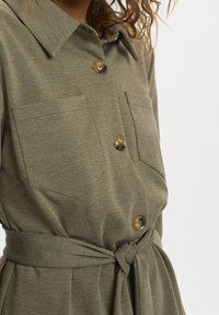 Olivenfarbenes Kleid mit kragenförmigem Ausschnitt, Fronttaschen und großen braunen Knöpfen. Verfügt über einen Bindegürtel für eine verstellbare Taille.