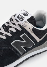 Primo piano di una sneaker New Balance 574 Classic nera e grigia con lacci bianchi, collo imbottito grigio e il distintivo logo "N" sul lato.