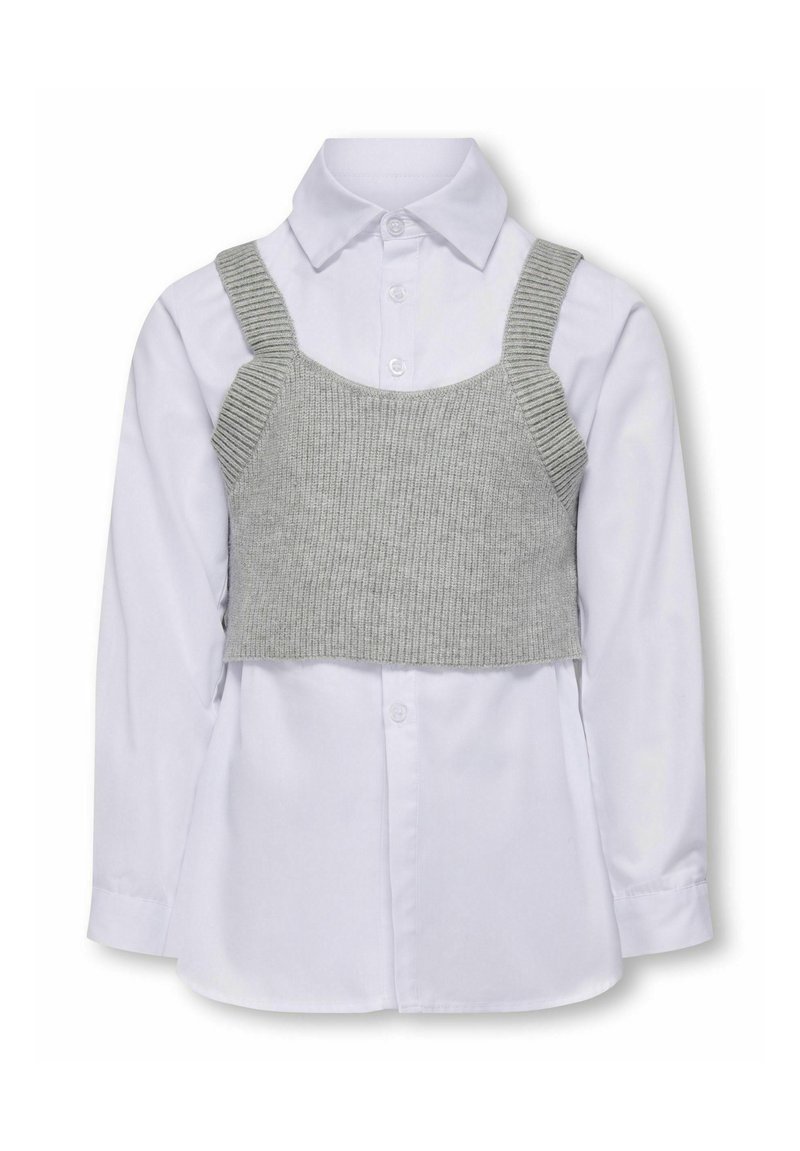 Grå strikket crop top over en hvid langærmet skjorte med knapper. Har stropper og ribbet tekstur på toppen. Knapp detalje på skjorten.