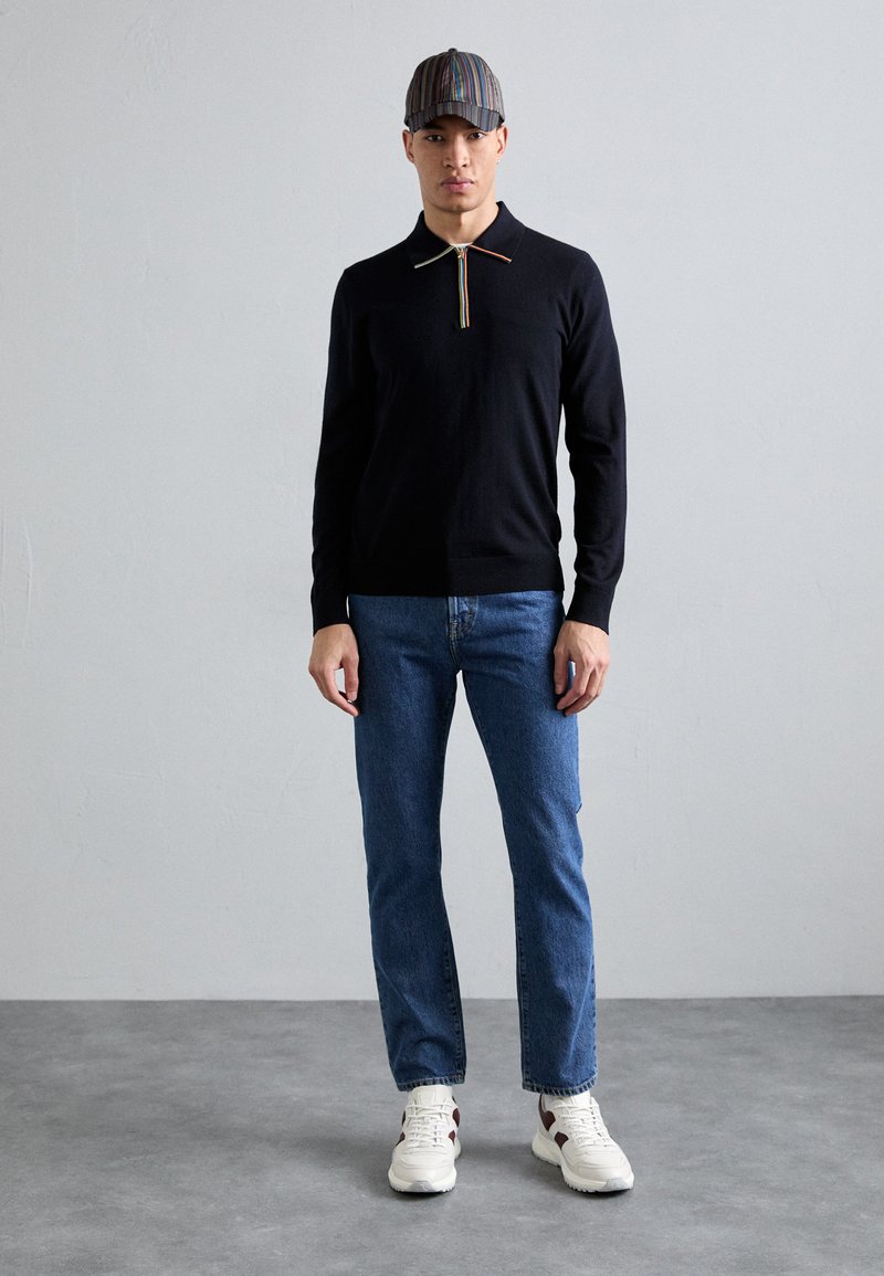 Ναυτικό πόλο μπλουζάκι με ουράνιο τόξο φερμουάρ, σε συνδυασμό με μπλε τζιν και λευκά αθλητικά παπούτσια. Το καπέλο έχει πολύχρωμες κατακόρυφες ρίγες.