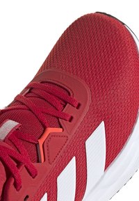 Red athletic shoe with textured mesh upper, contrasting white stripes, and orange accents. Features lace-up design and padded collar.