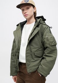 Olīvzaļš pūšamais mētelis ar rāvējslēdzēju priekšpusē, vairākiem kabatām, regulējamu kapuci un melnu oderi, uzvilkts virs baltas krekla un brūnām biksēm.