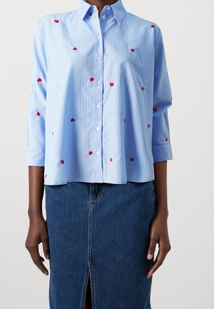 Chemise à rayures bleu clair avec petits cœurs rouges brodés, portée avec une jupe en denim bleu foncé à fente avant et fermeture à boutons.